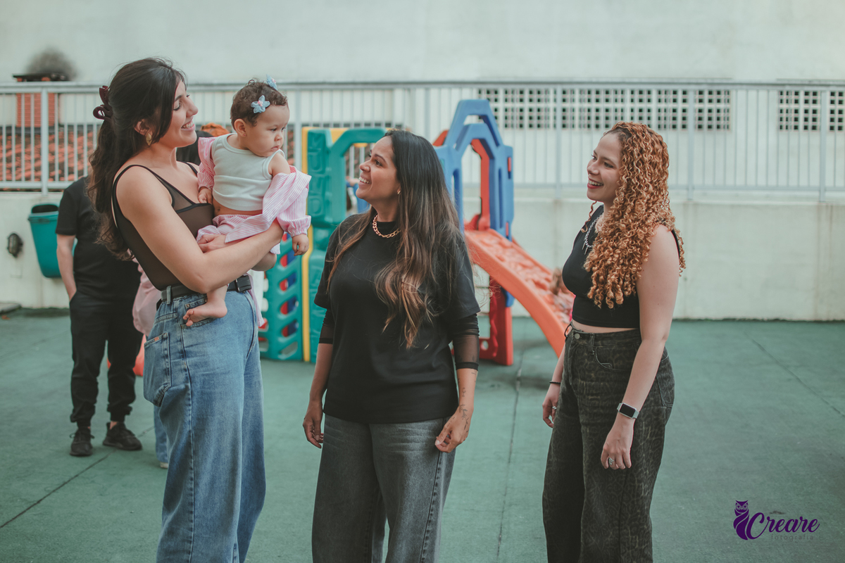Uma mulher sorrindo com uma bebê no colo e várias pessoas ao lado sorrindo, durante festa de aniversário em casa.