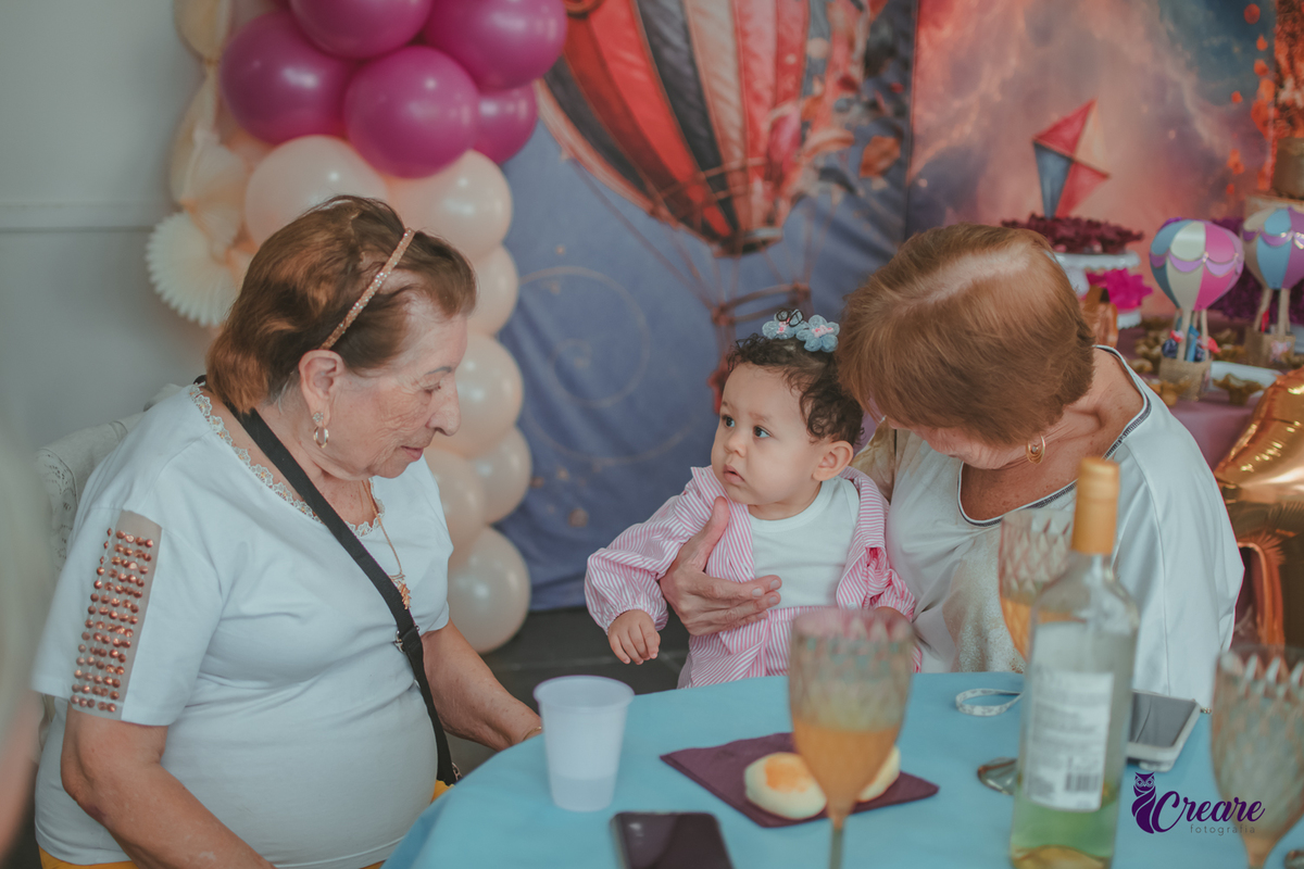 Duas senhoras com uma bebê de 1 ano no colo durante festa de aniversário em casa.