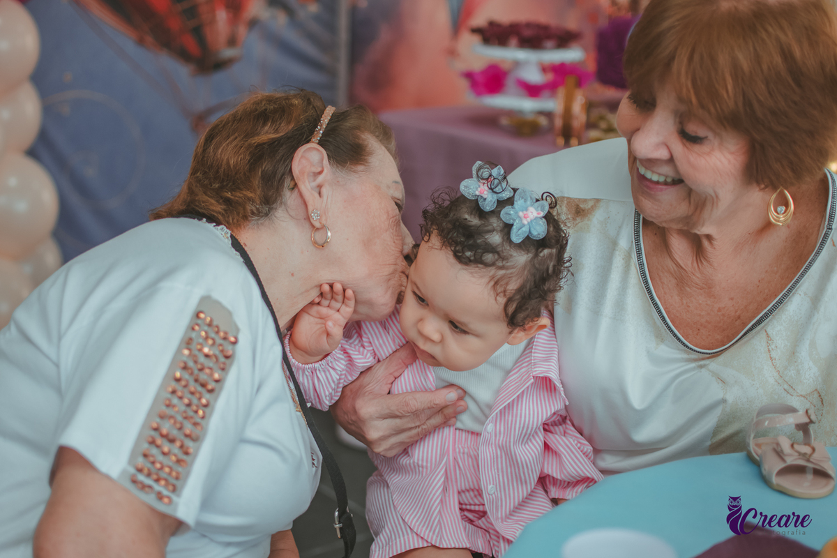 Uma senhora dando beijo no rosto de uma bebê que está no colo da sua avó, durante festa de aniversário de 1 ano em casa.