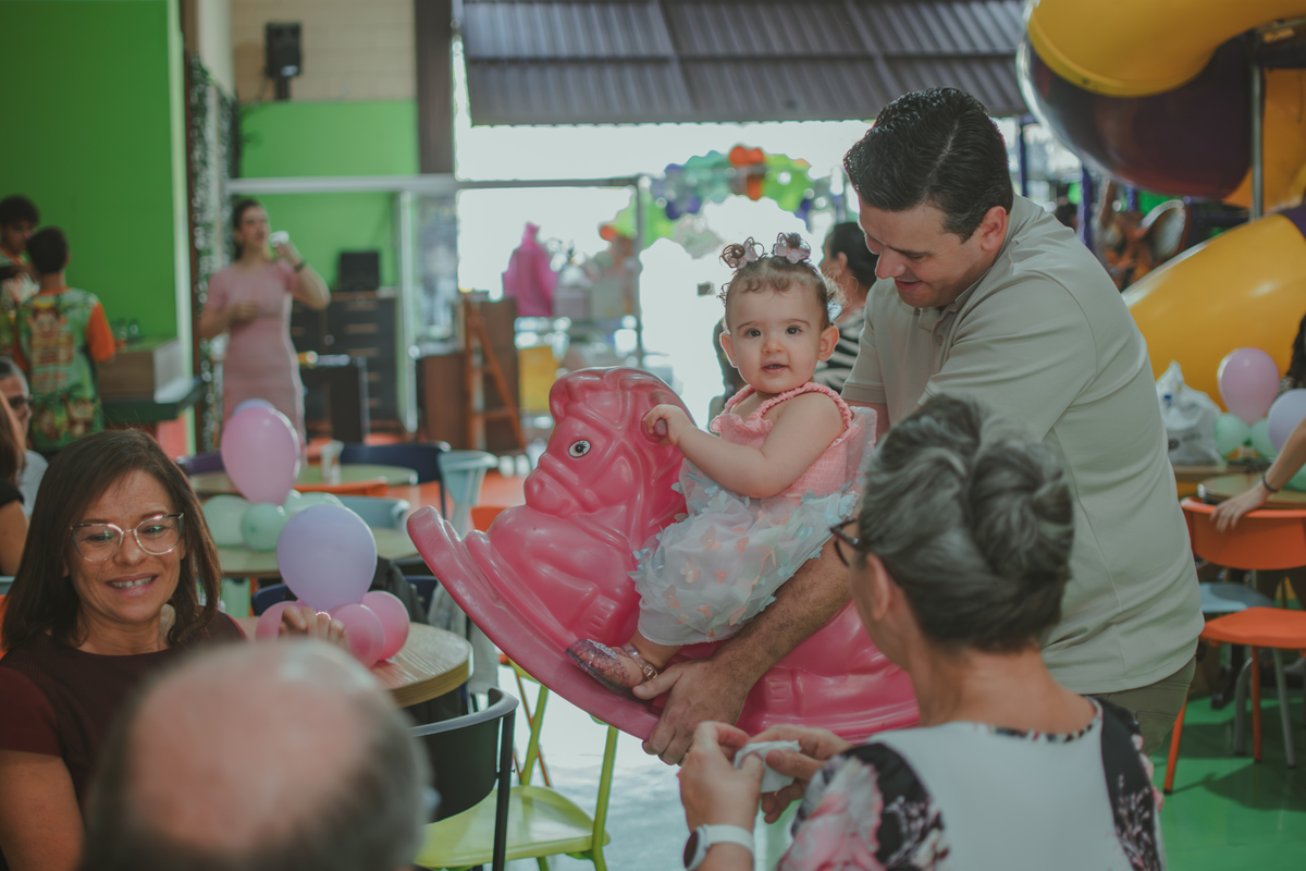 Uma homem carregando uma bebê em um brinquedo em formato de cavalinho rosa, durante festa de aniversário infantil.