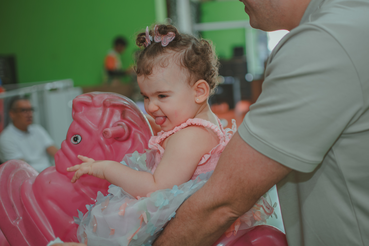 Uma homem carregando uma bebê em um brinquedo em formato de cavalinho rosa, durante festa de aniversário infantil.