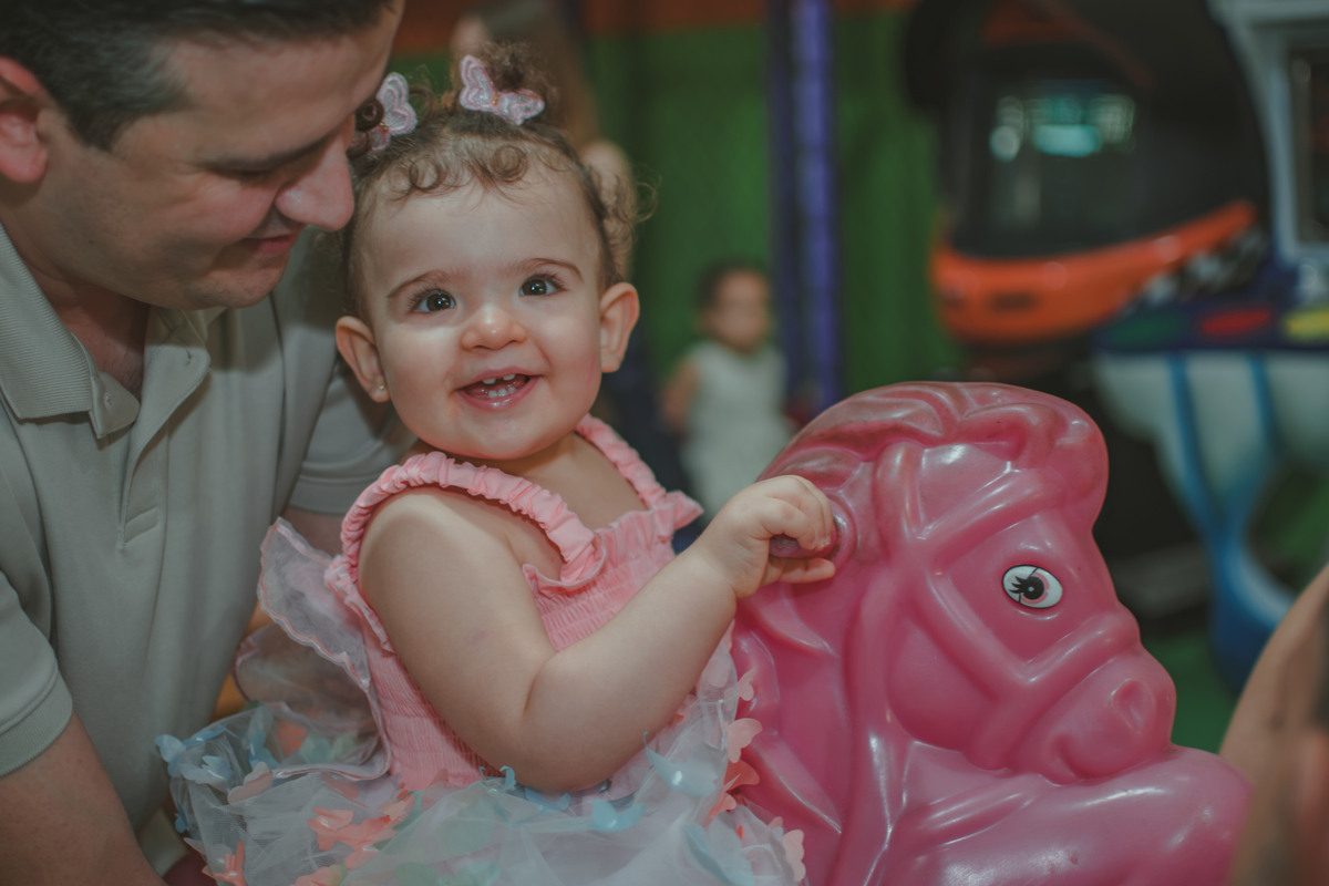 Uma homem carregando uma bebê em um brinquedo em formato de cavalinho rosa, durante festa de aniversário infantil.