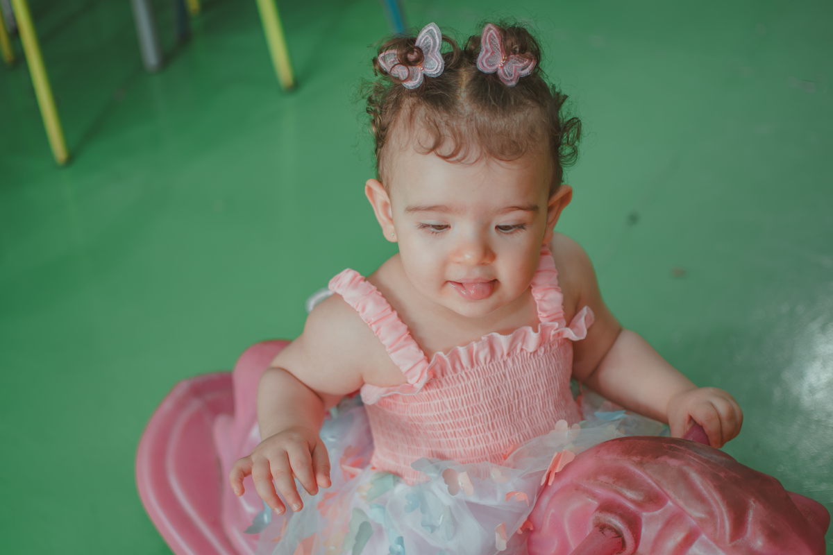 Uma bebê em um brinquedo em formato de cavalinho rosa, durante festa de aniversário infantil.