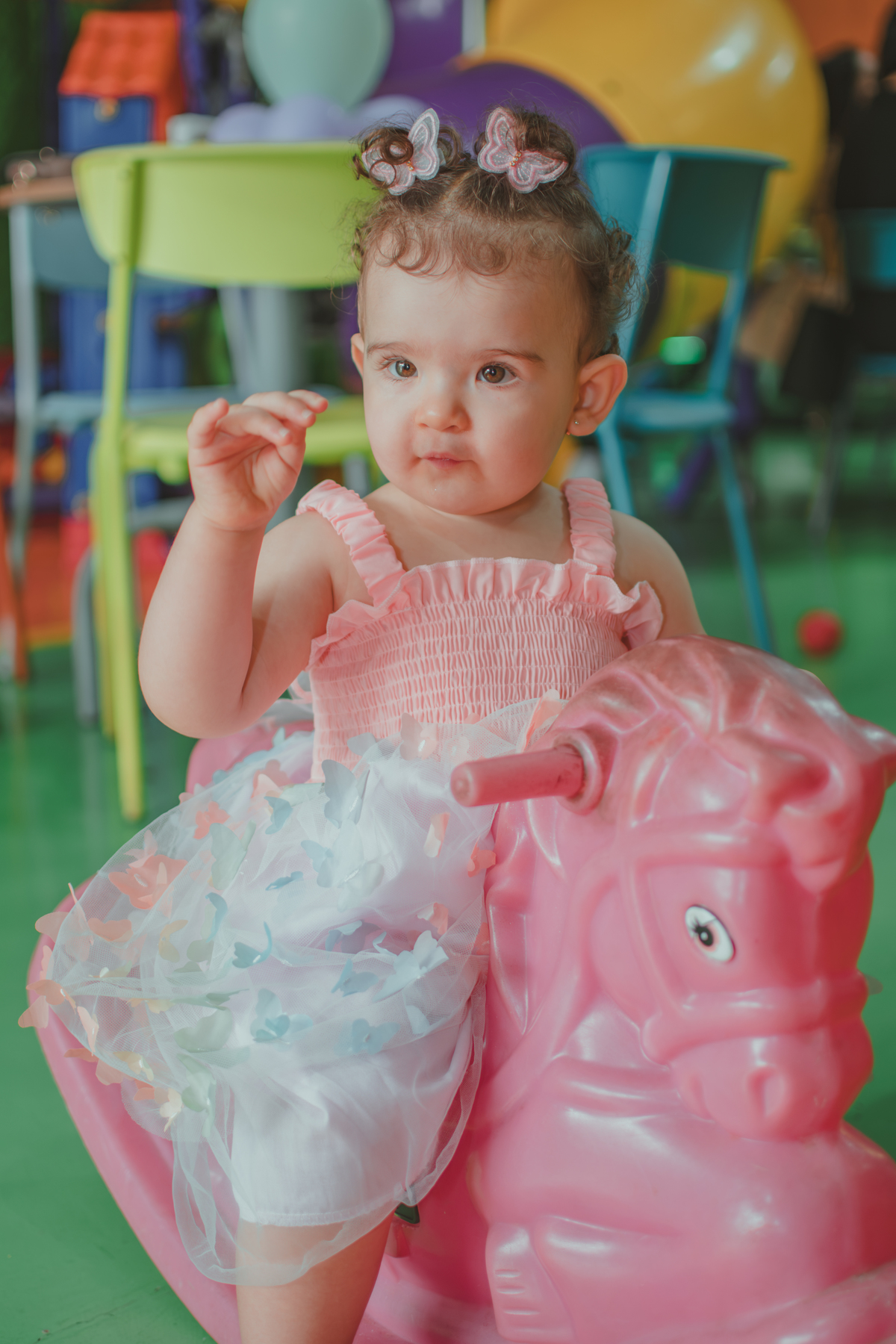 Uma bebê em um brinquedo em formato de cavalinho rosa, durante festa de aniversário infantil.
