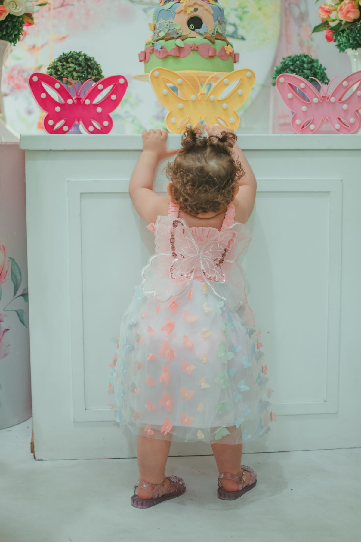 Uma bebê de costas, mostrando os detalhes do seu vestido que tem uma asa de borboleta.