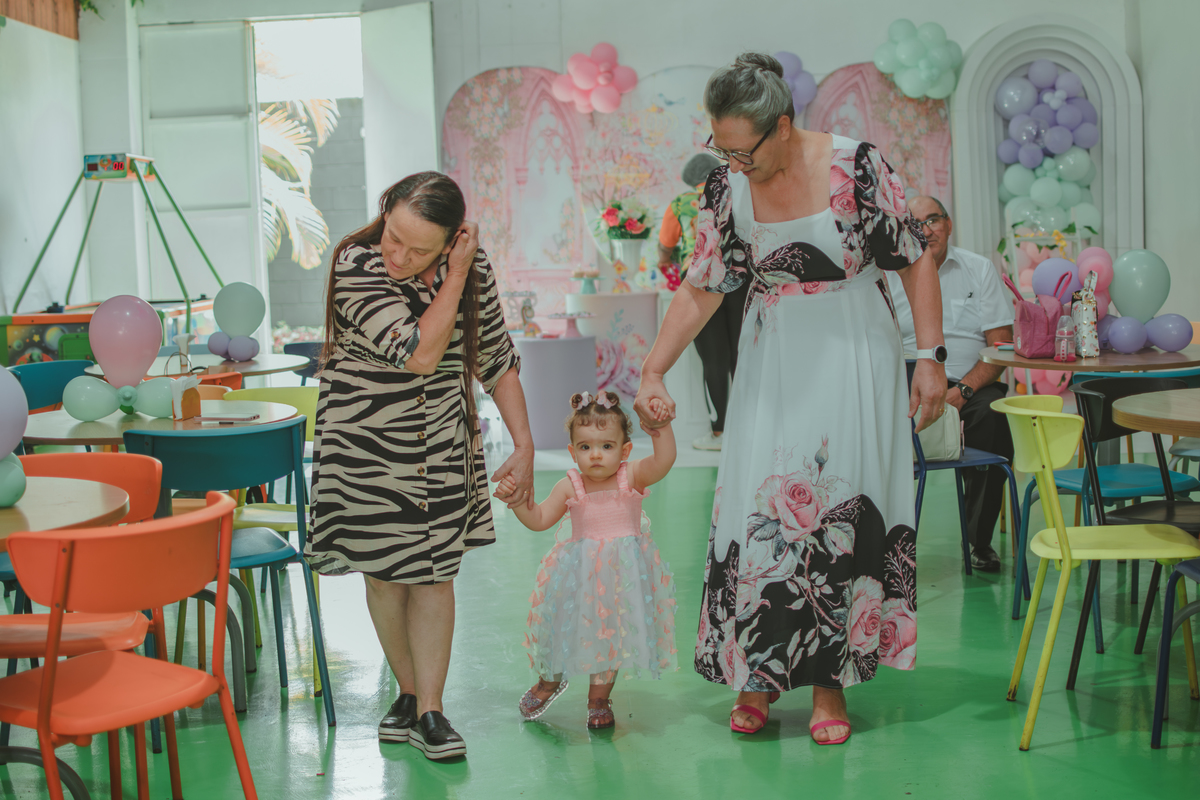 Duas senhoras segurandona mão de uma bebê que caminha pelo salão da sua festa de aniversário de 1 ano.