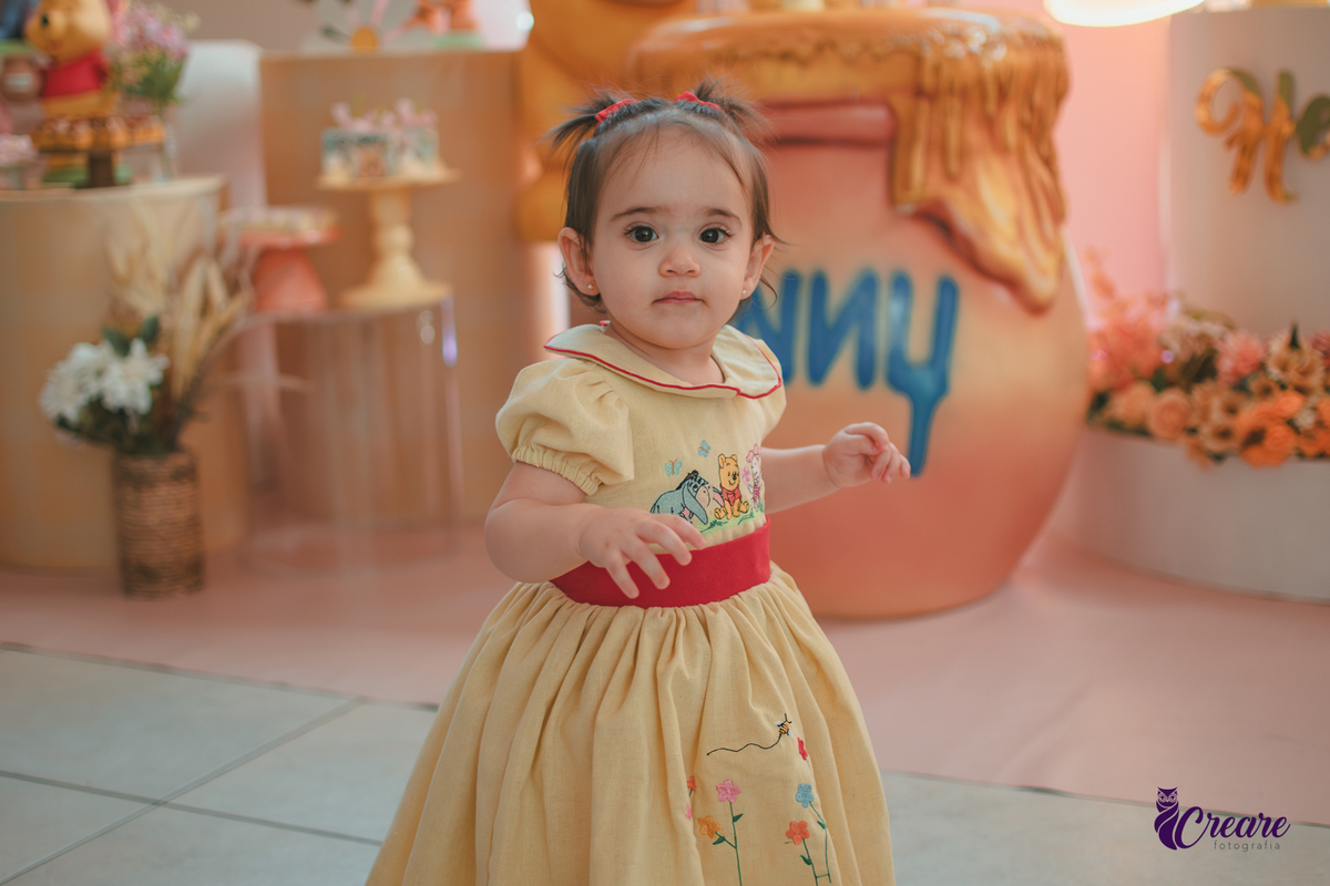 Uma bebê, usando vestido amarelo com faixa vermelha e andando pelo salão da festa de seu aniversário de 1 ano, feita em casa.