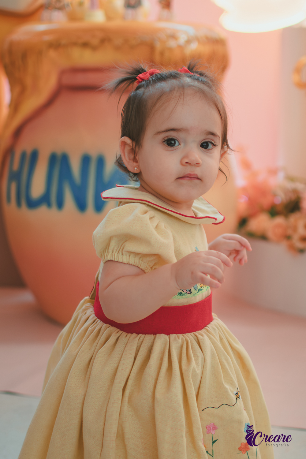 Uma bebê, usando vestido amarelo com faixa vermelha e andando pelo salão da festa de seu aniversário de 1 ano, feita em casa.