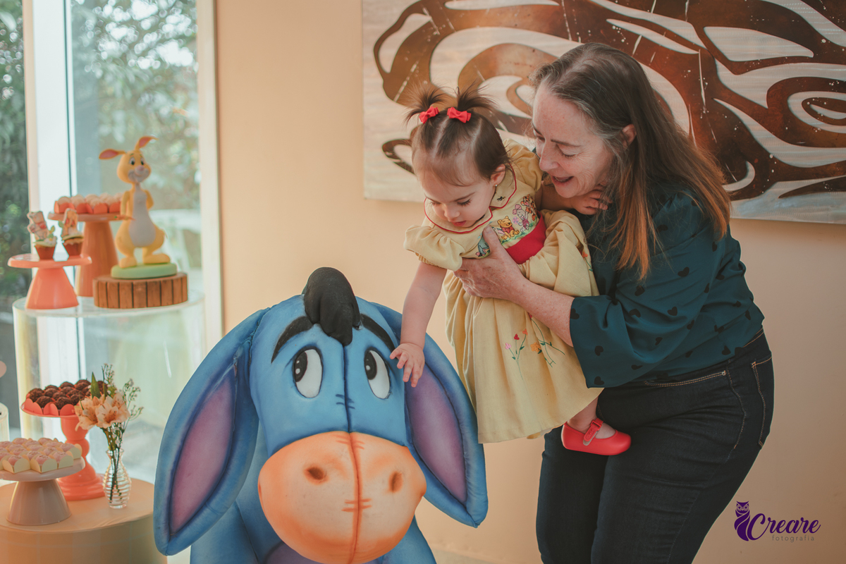 Uma mulher segurando uma bebê no colo enquanto brincam com um boneco gigante do Bizonho, personagem do ursinho Pooh, tema da festa de aniversário feita em casa.