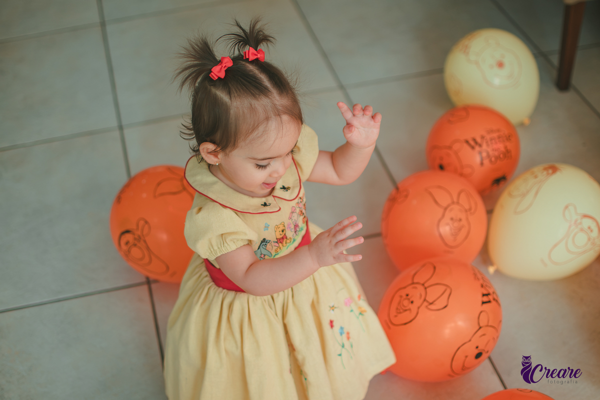 Uma bebê brincando entre balões amarelos e laranjaas, durante sua festa de aniversário de 1 ano em casa.