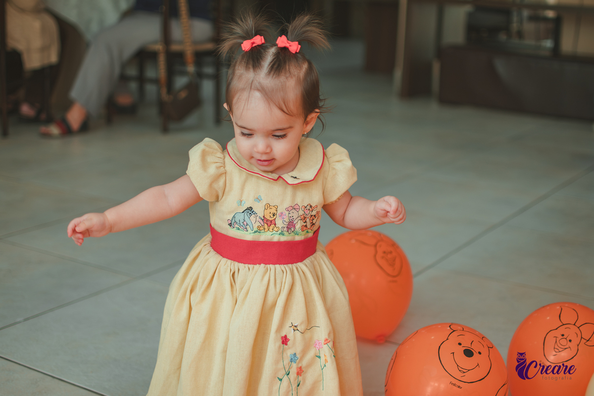 Uma bebê brincando entre balões amarelos e laranjaas, durante sua festa de aniversário de 1 ano em casa.