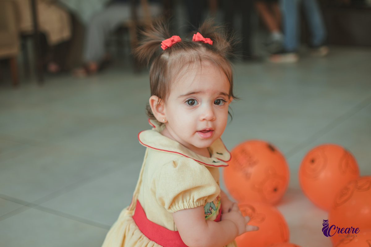Uma bebê brincando entre balões amarelos e laranjaas, durante sua festa de aniversário de 1 ano em casa.