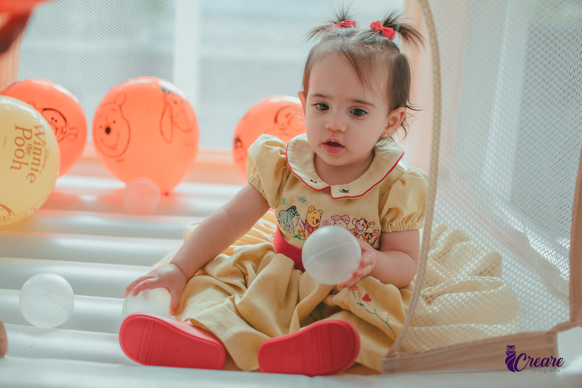 Uma bebê brincando no brinquedo inflável, cercada por balões laranjas e bolinhas brancas, durante sua festa de aniversário de 1 ano.