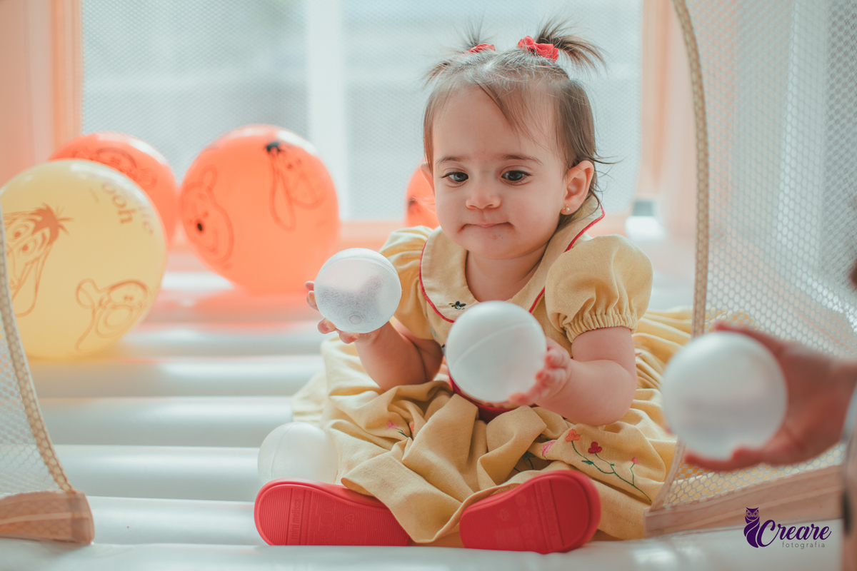 Uma bebê brincando no brinquedo inflável, cercada por balões laranjas e bolinhas brancas, durante sua festa de aniversário de 1 ano.