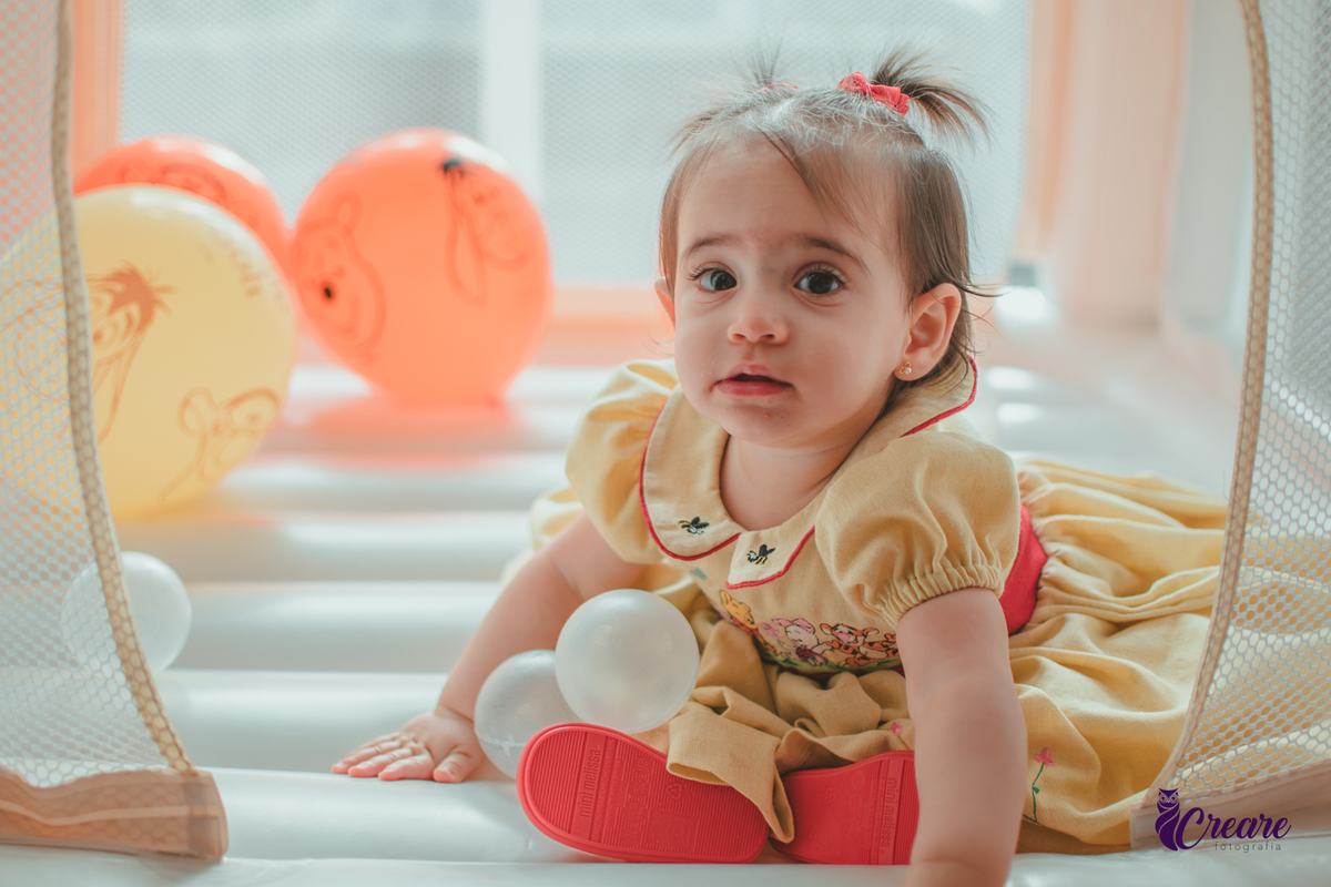 Uma bebê brincando no brinquedo inflável, cercada por balões laranjas e bolinhas brancas, durante sua festa de aniversário de 1 ano.