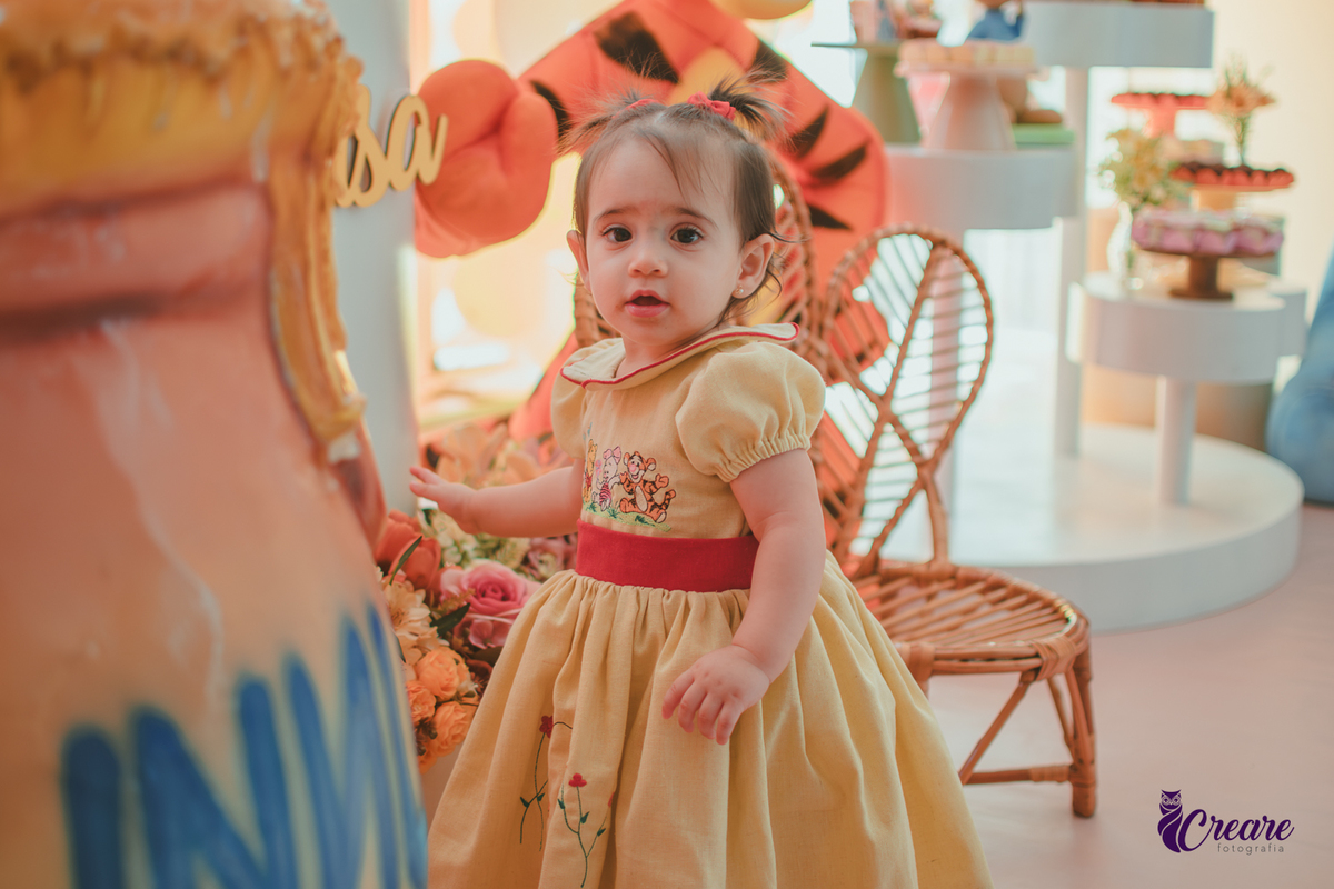 Uma bebê, usando vestido amarelo com faixa vermelha e andando pelo salão da festa de seu aniversário de 1 ano, feita em casa.
