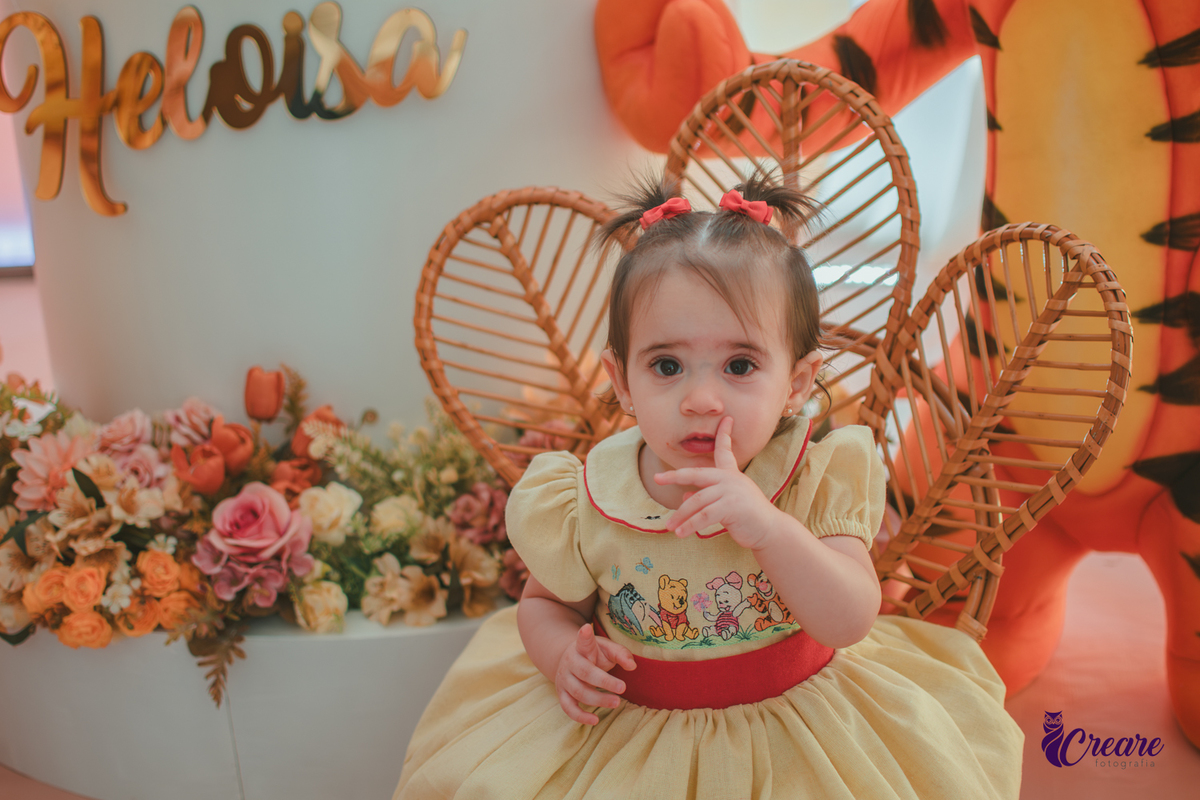 Uma bebê, usando vestido amarelo com faixa vermelha e sentada em frente a decoração de seu aniversário de 1 ano, feita em casa.