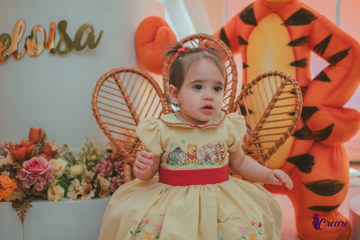 Uma bebê, usando vestido amarelo com faixa vermelha e sentada em frente a decoração de seu aniversário de 1 ano, feita em casa.