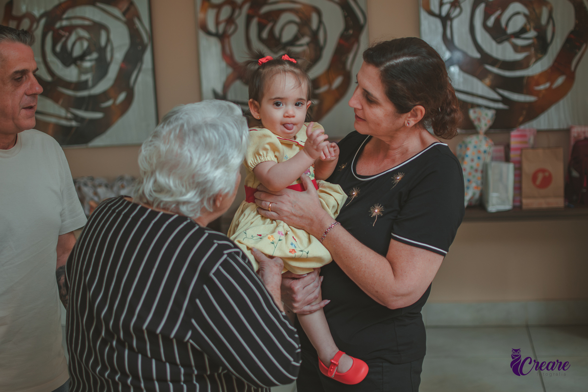 Duas mulheres, interagindo com uma bebê de 1 ano, durante festa de aniversário em casa.