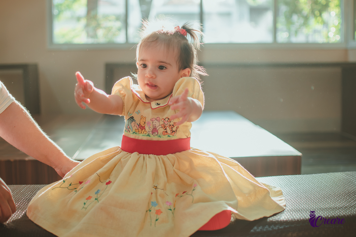 Uma bebê, brincando com reflexos de sol atrás dela, durante sua festa de aniversário de 1 ano feita em casa.