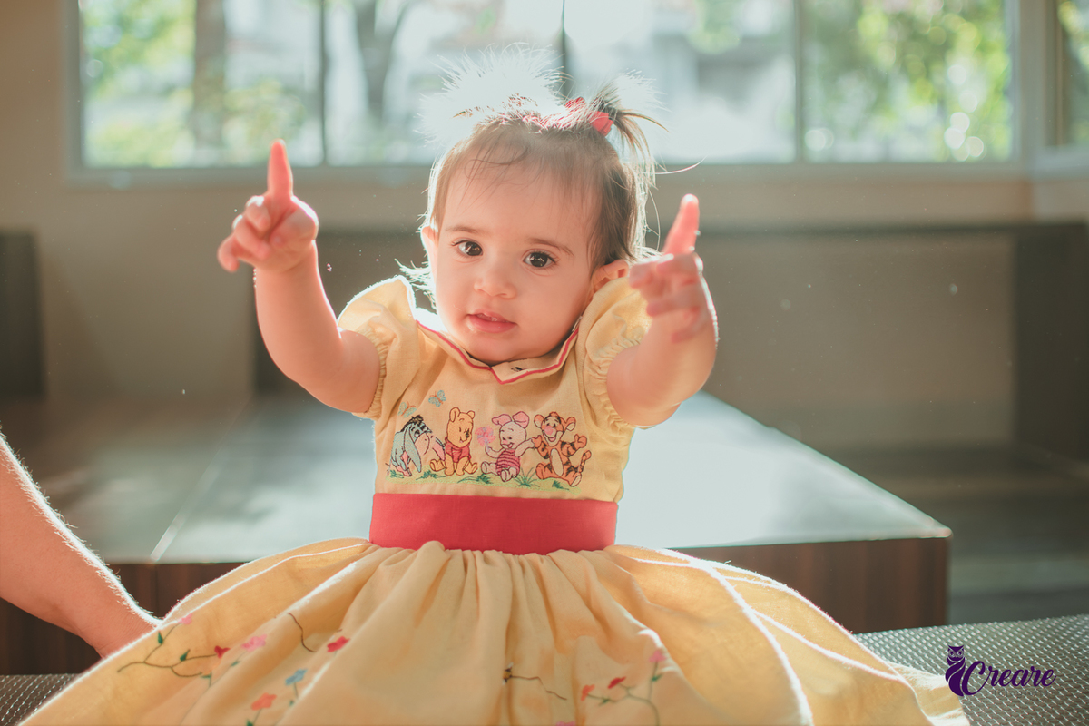 Uma bebê, brincando com reflexos de sol atrás dela, durante sua festa de aniversário de 1 ano feita em casa.
