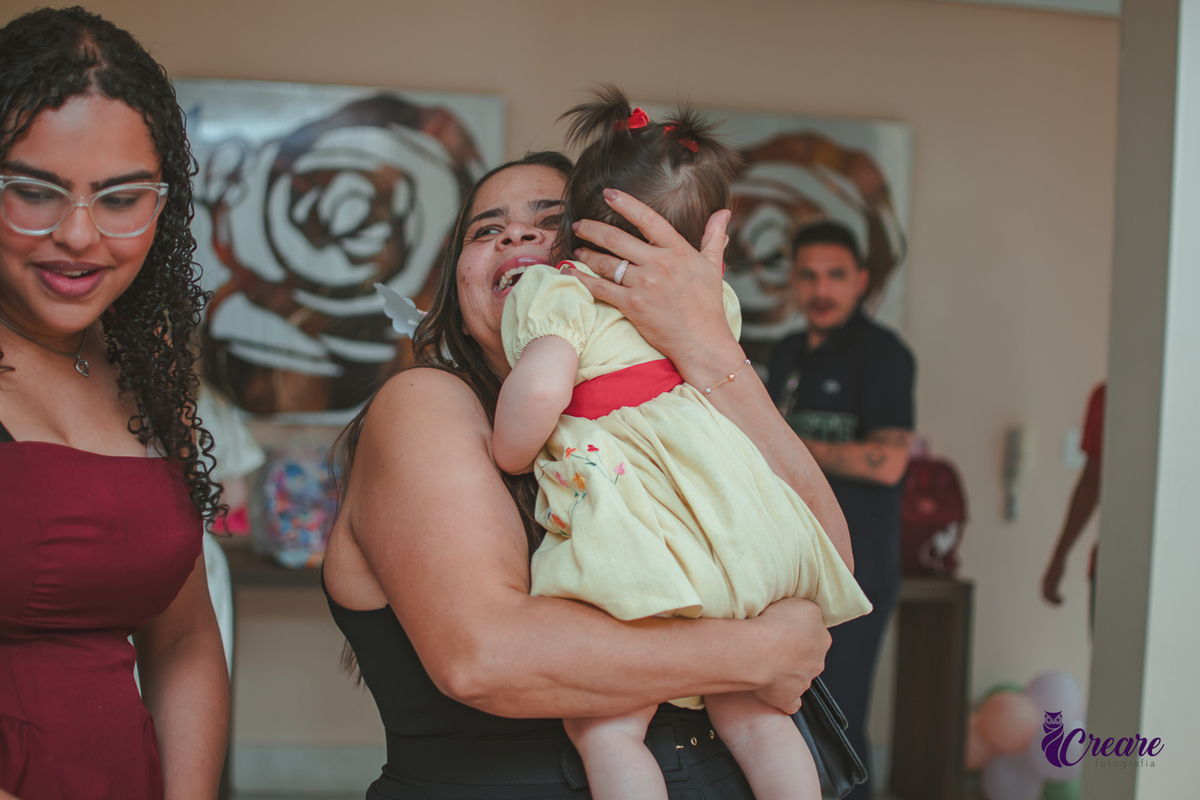 Uma mulher abraçando uma bebê e sorrindo, durante festa de 1 ano em casa.