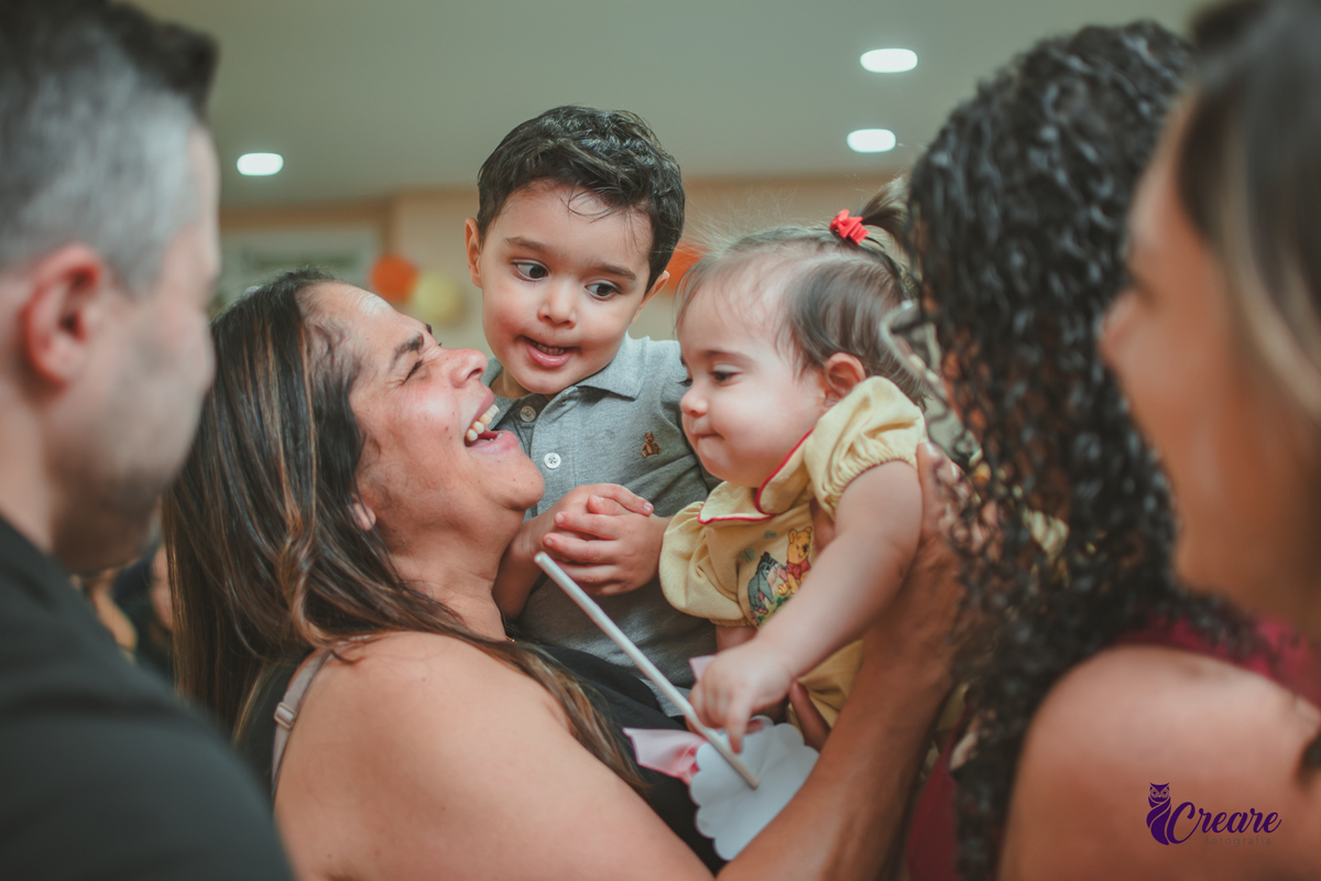 Uma mulher abraçando dois bebês e sorrindo, durante festa de 1 ano em casa.