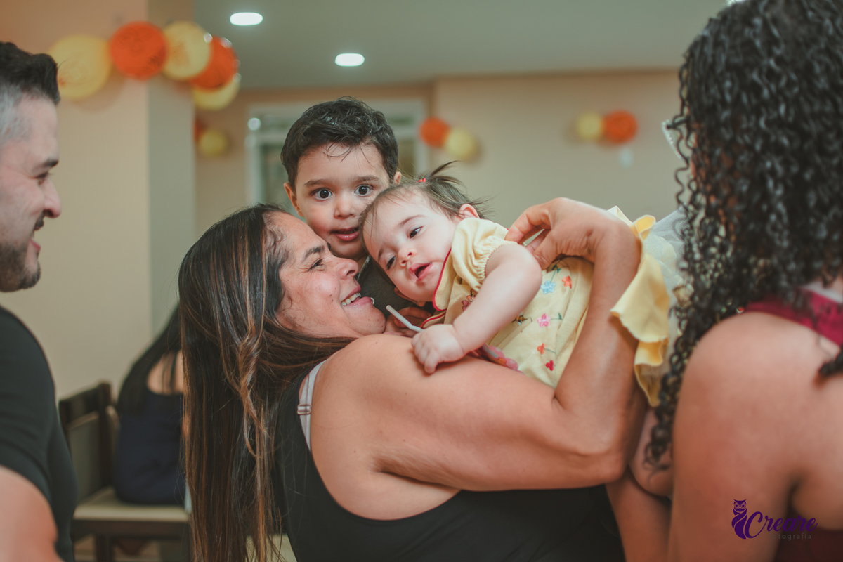 Uma mulher abraçando dois bebês e sorrindo, durante festa de 1 ano em casa.