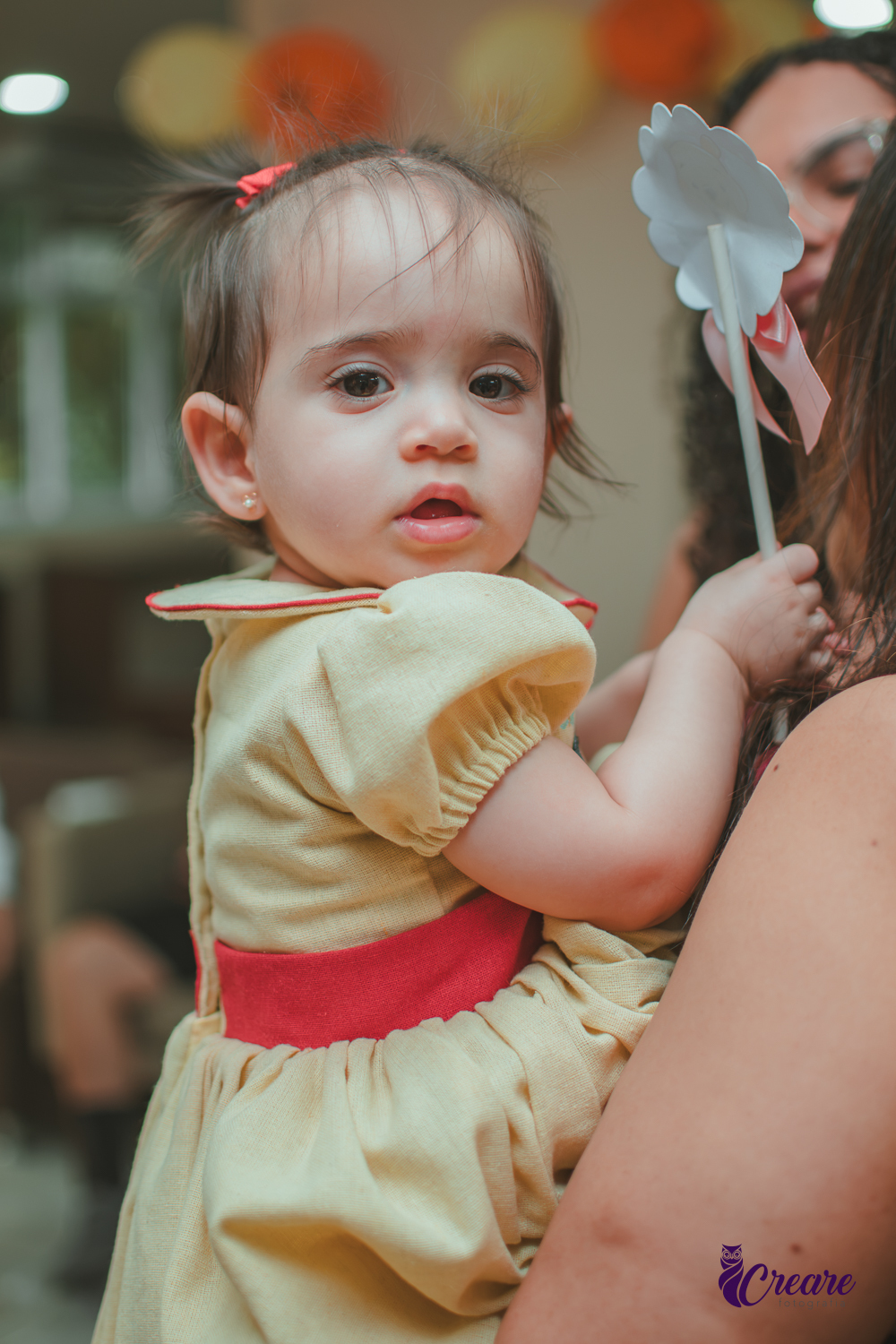 Uma bebê no colo de uma mulher e olhando fixamente para frente, durante festa de aniversário de 1 ano.