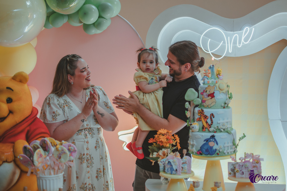 Um casal, em uma decoração de ursinho Pooh, cantando parabéns, durante festa de aniversário de 1 ano.