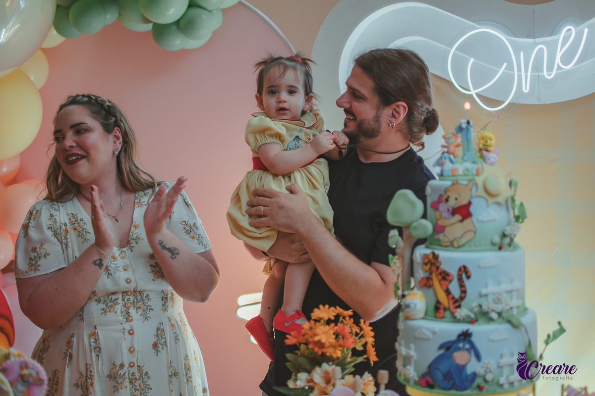 Um casal, em uma decoração de ursinho Pooh, cantando parabéns, durante festa de aniversário de 1 ano.