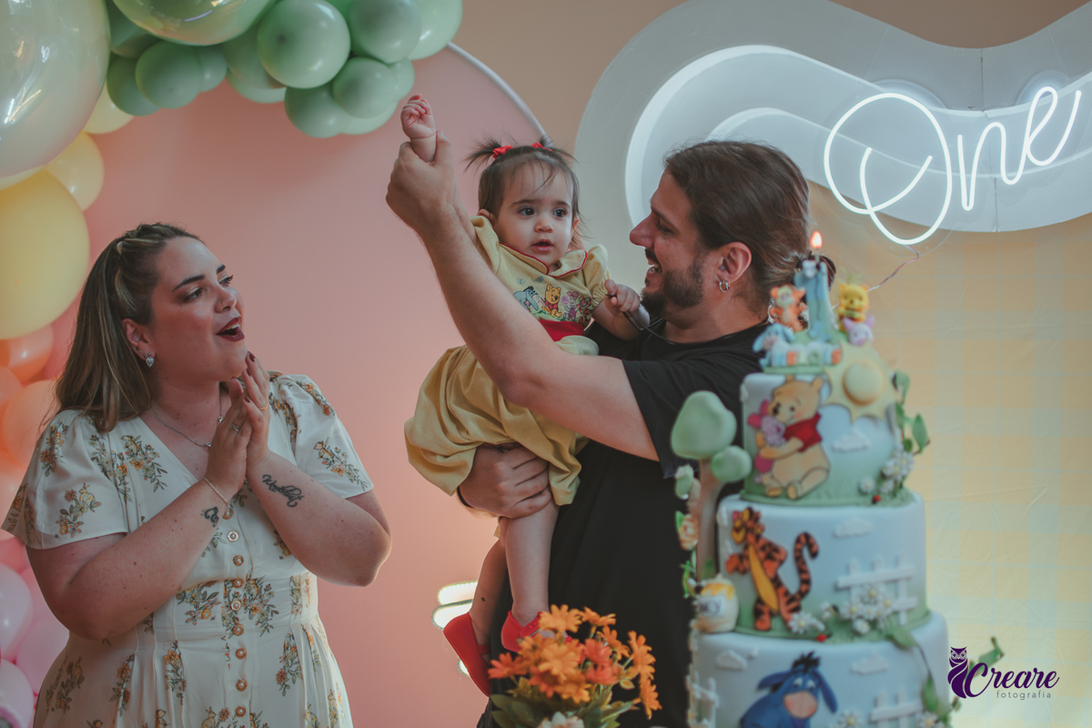 Um casal, em uma decoração de ursinho Pooh, cantando parabéns, durante festa de aniversário de 1 ano.