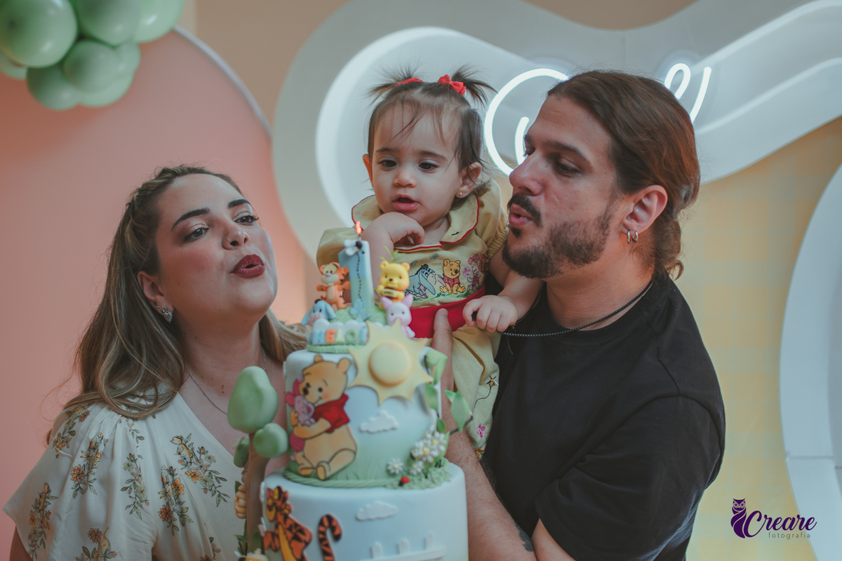 Um casal, em uma decoração de ursinho Pooh, soprando velinha de parabéns, durante festa de aniversário de 1 ano.