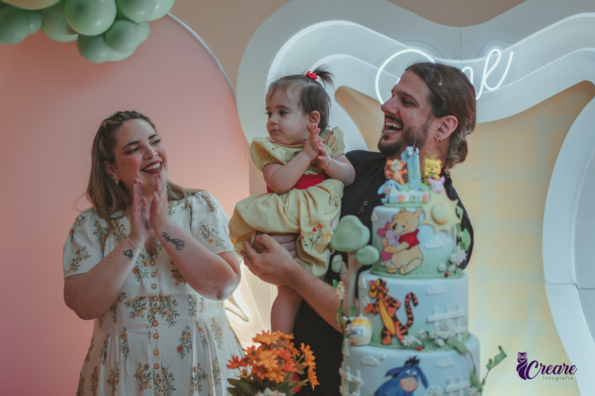 Um casal, em uma decoração de ursinho Pooh, cantando parabéns, durante festa de aniversário de 1 ano.