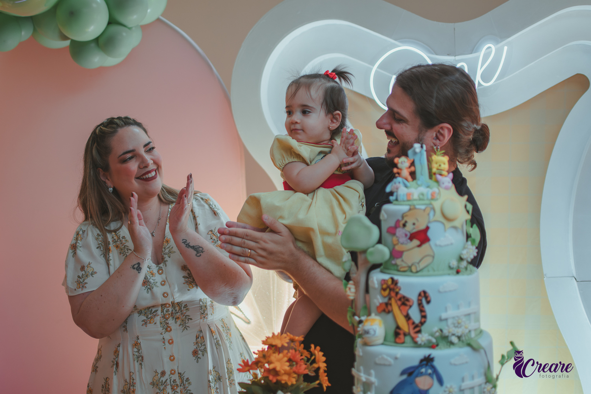 Um casal, em uma decoração de ursinho Pooh, cantando parabéns, durante festa de aniversário de 1 ano.