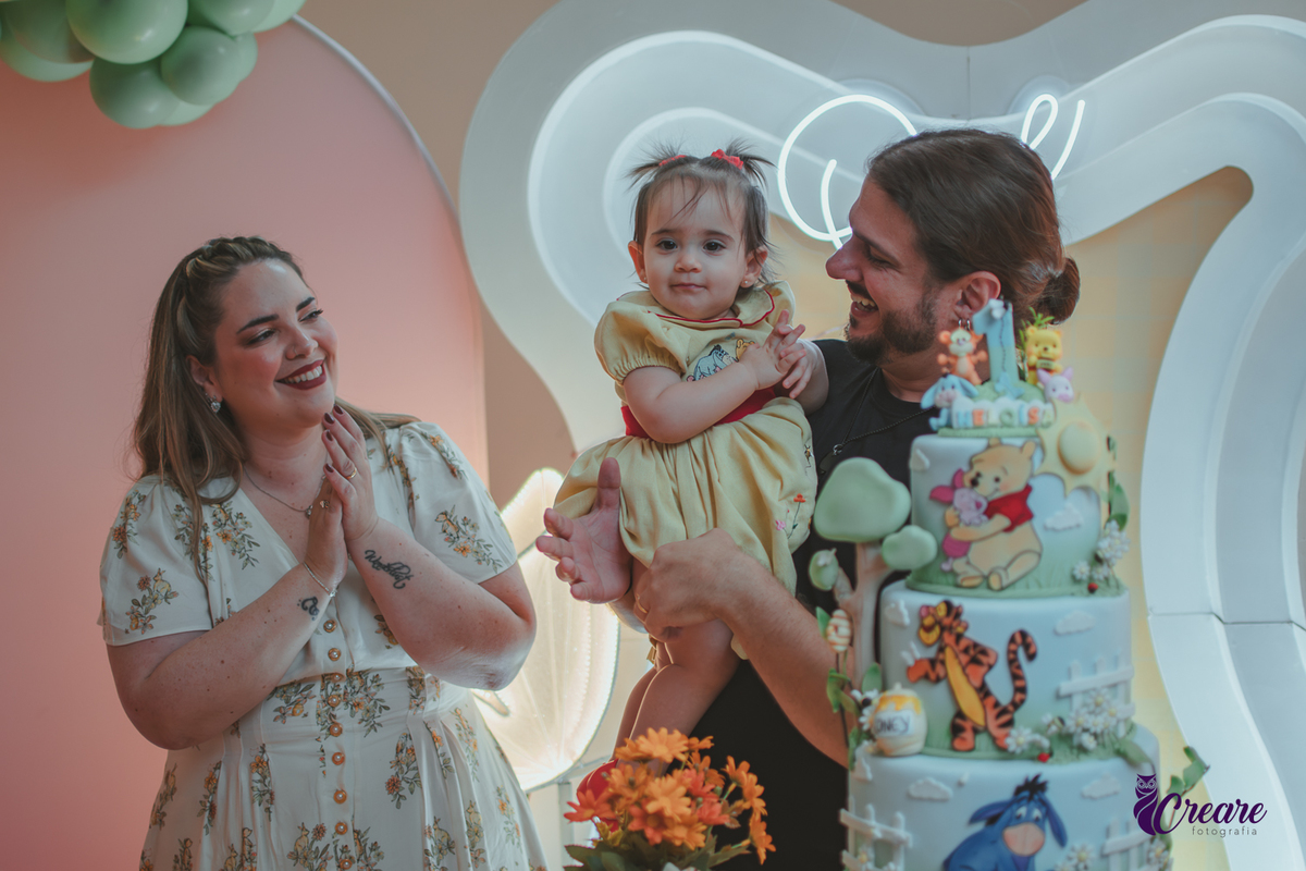 Um casal, em uma decoração de ursinho Pooh, cantando parabéns, durante festa de aniversário de 1 ano.