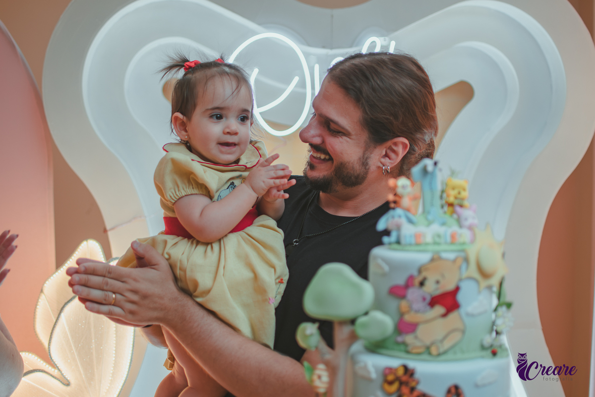 Um homem, em uma decoração de ursinho Pooh, cantando parabéns, durante festa de aniversário de 1 ano.