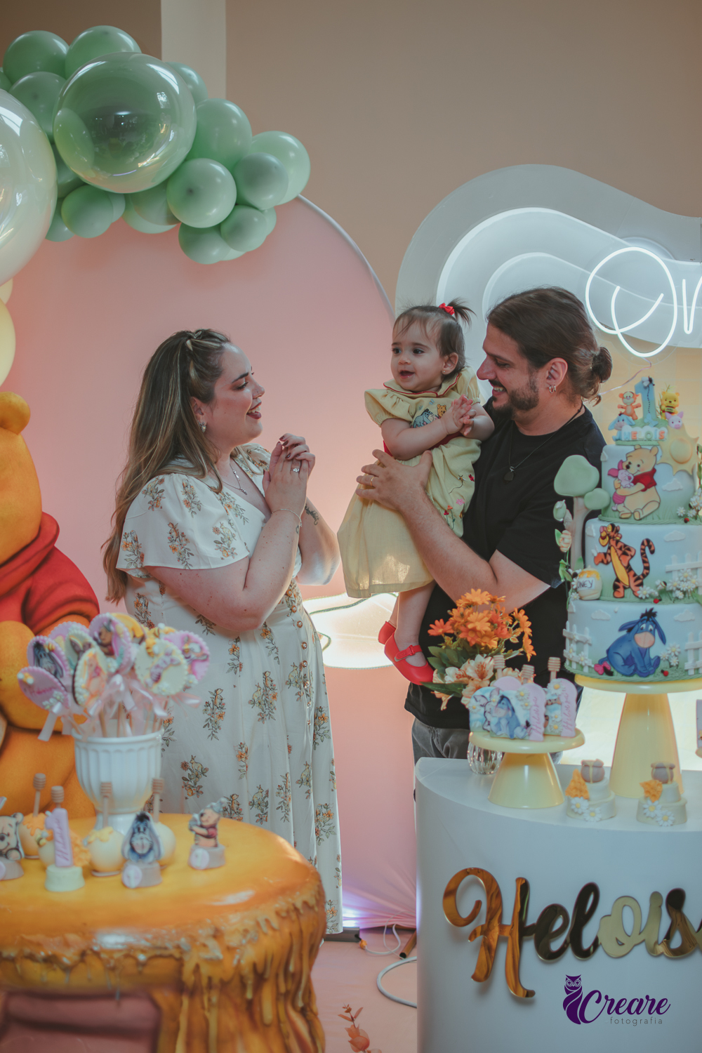 Um casal, em uma decoração de ursinho Pooh, cantando parabéns, durante festa de aniversário de 1 ano.