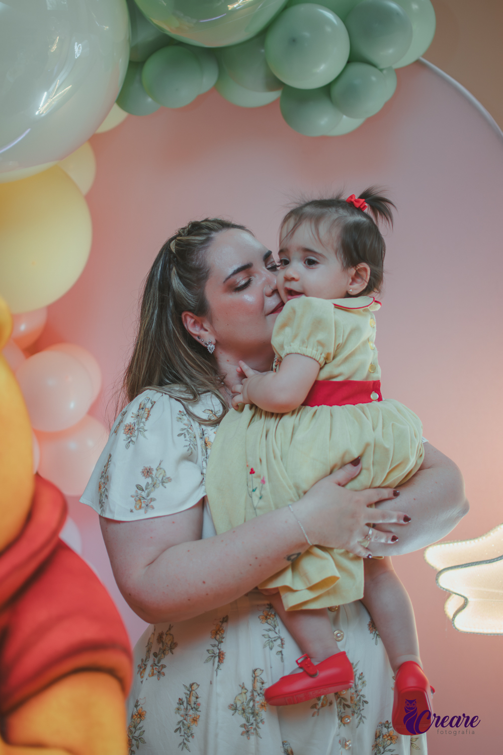Uma mulher, em uma decoração de ursinho Pooh, beijando o rosto da sua filha, durante festa de aniversário de 1 ano.