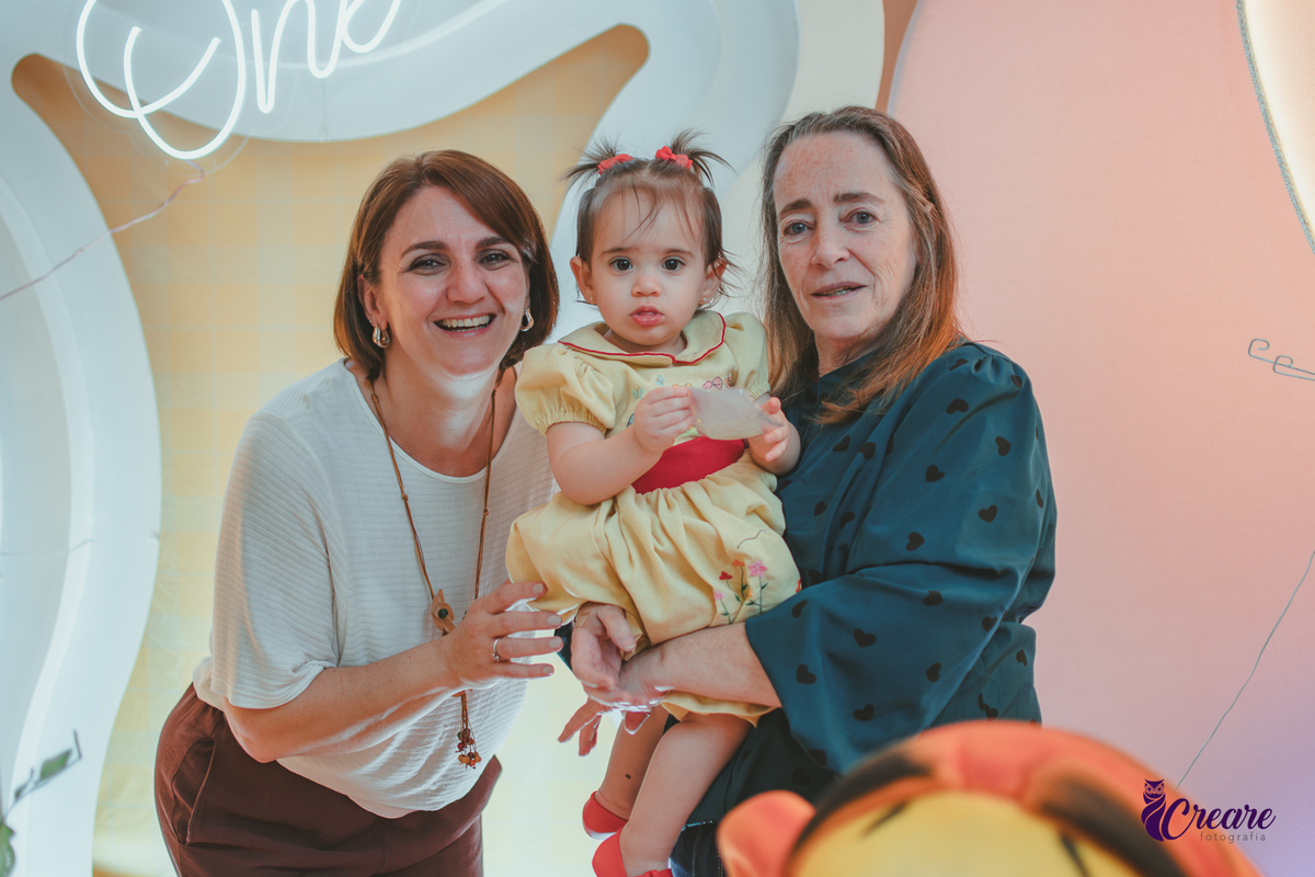 Duas mulheres, segurando uma bebê no colo em frente uma mesa de decoração de aniversário infantil.