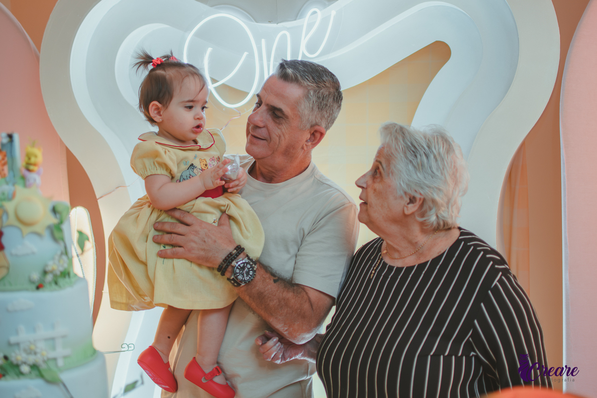 Um homem e uma senhora de idade, segurando uma bebê no colo em frente uma mesa de decoração de aniversário infantil.