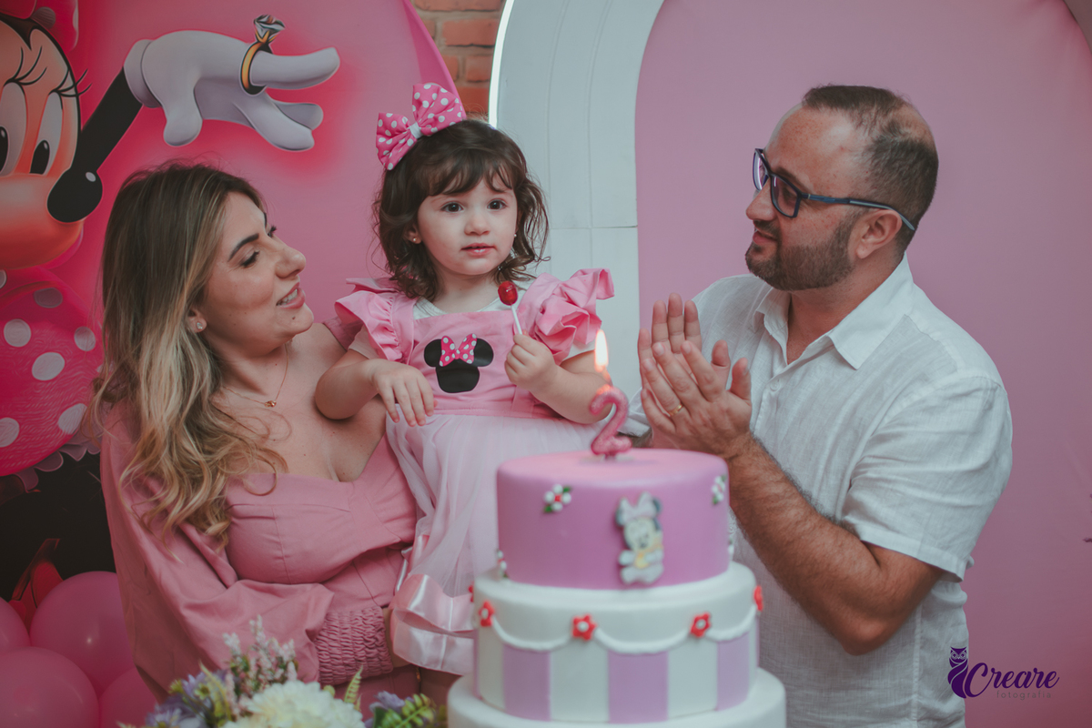 uma familia em frente uma mesa de decoração com tema Minnie, cantando parabéns durante festa de aniversário infantil.