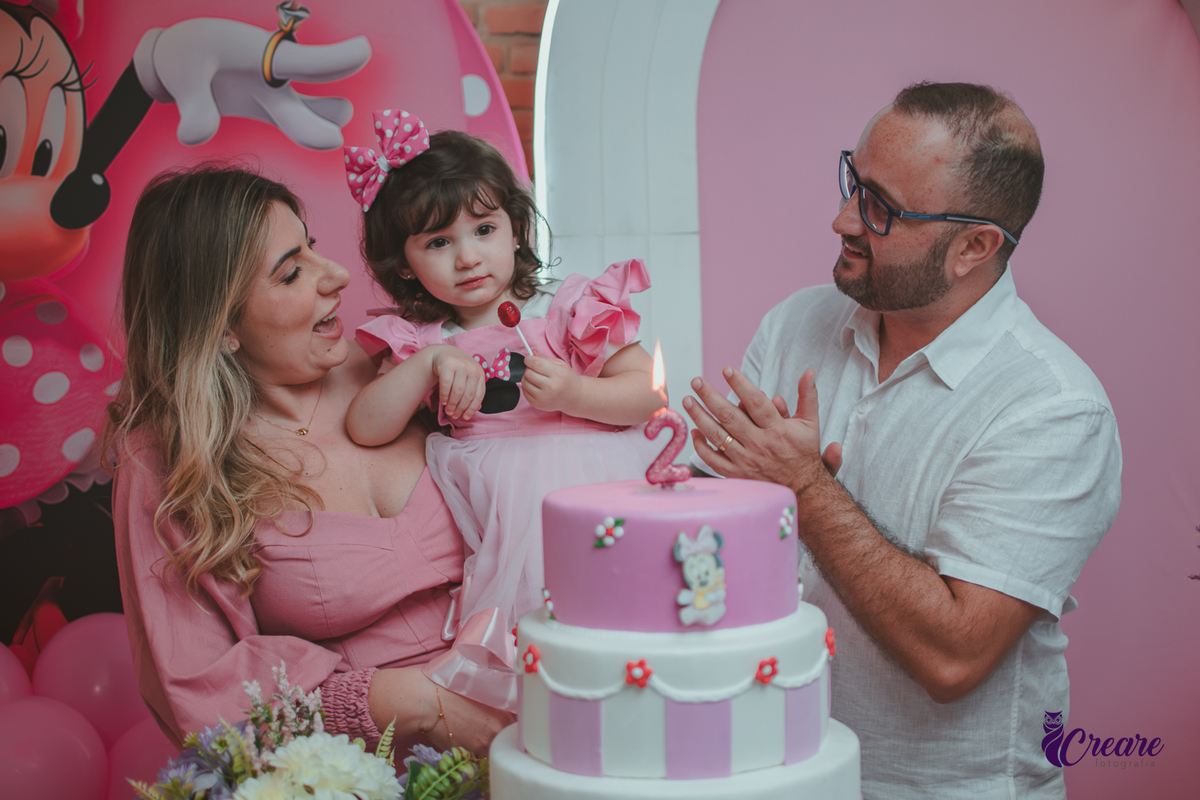 uma familia em frente uma mesa de decoração com tema Minnie, cantando parabéns durante festa de aniversário infantil.