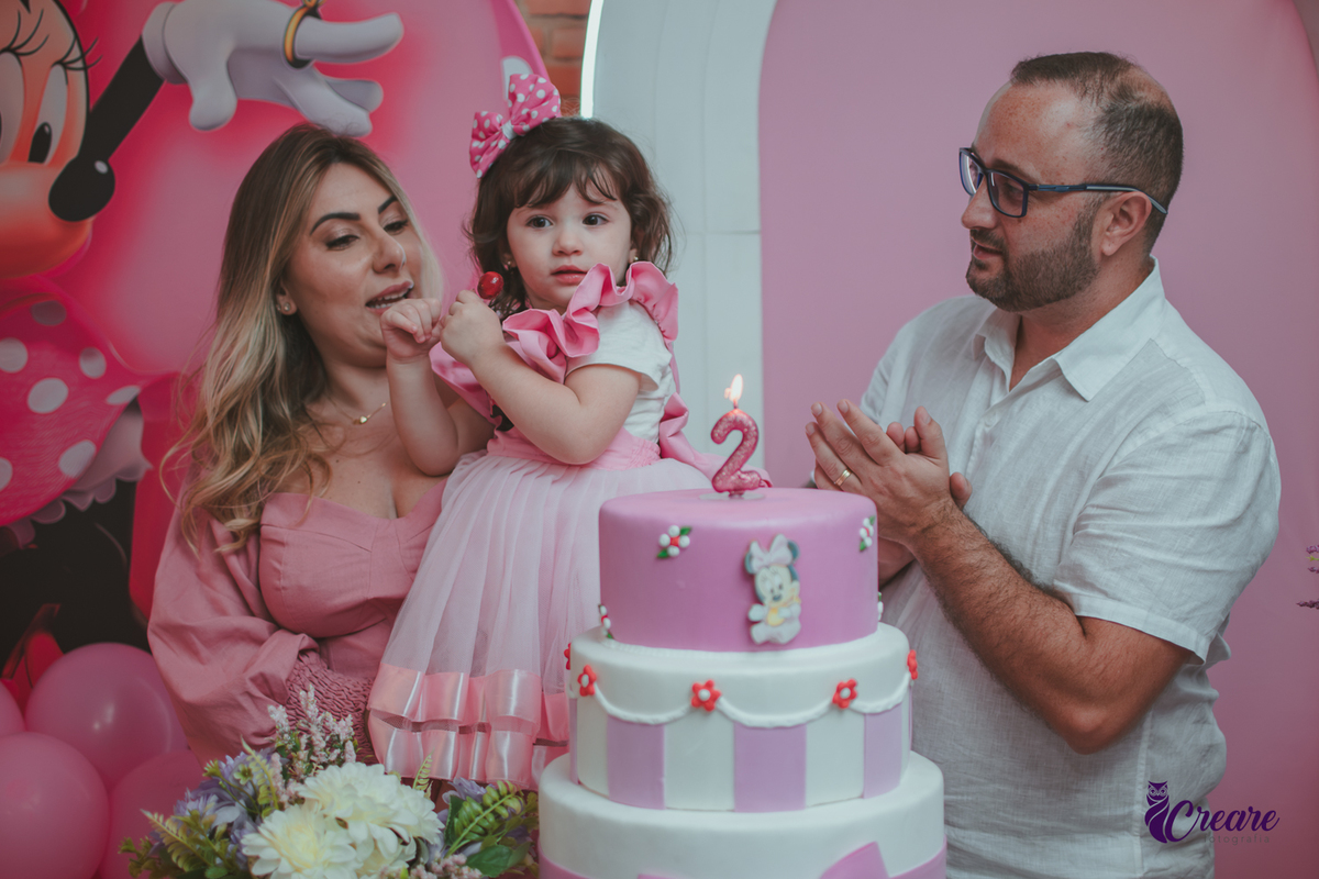 uma familia em frente uma mesa de decoração com tema Minnie, cantando parabéns durante festa de aniversário infantil.