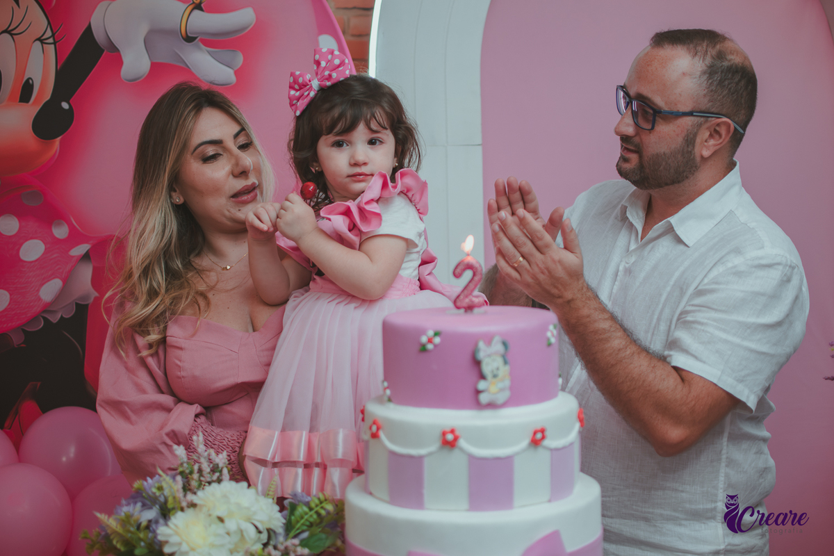 uma familia em frente uma mesa de decoração com tema Minnie, cantando parabéns durante festa de aniversário infantil.