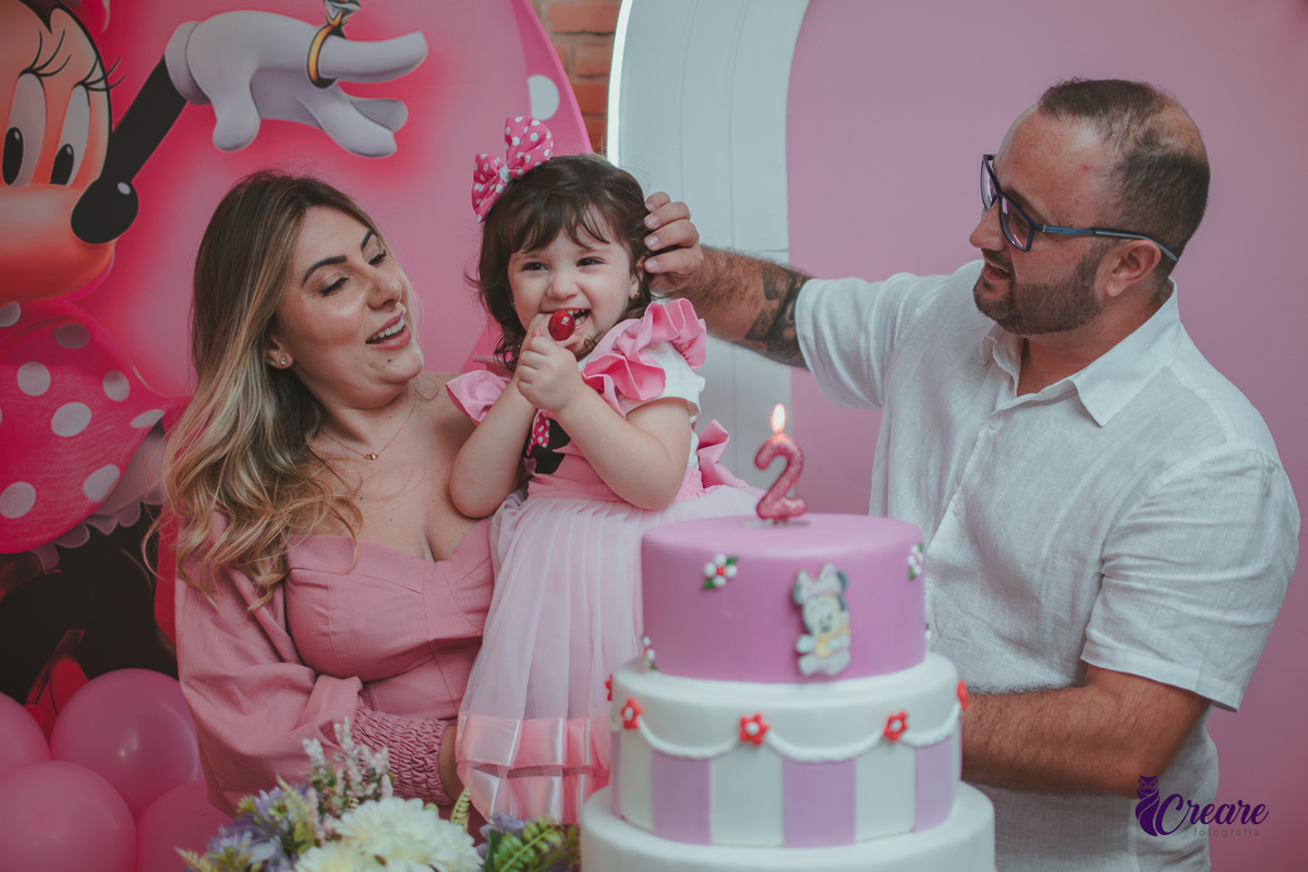 uma familia em frente uma mesa de decoração com tema Minnie, cantando parabéns durante festa de aniversário infantil.