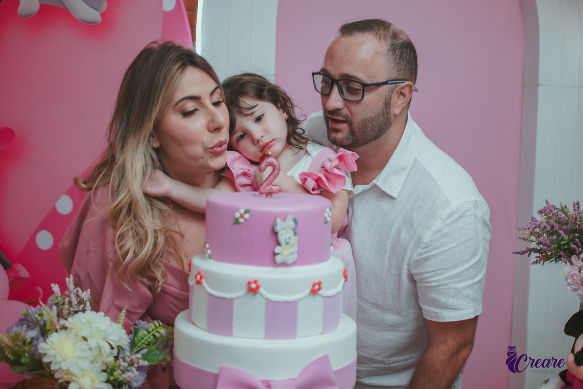 uma familia em frente uma mesa de decoração com tema Minnie, cantando parabéns durante festa de aniversário infantil.