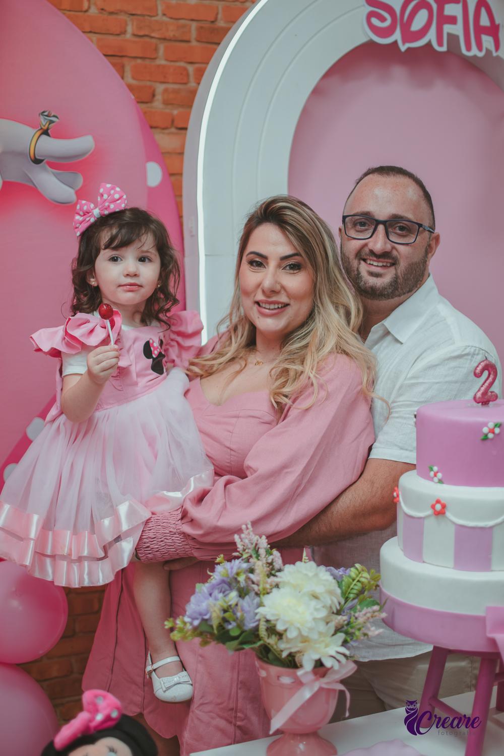 uma familia em frente uma mesa de decoração com tema Minnie, durante festa de aniversário infantil.