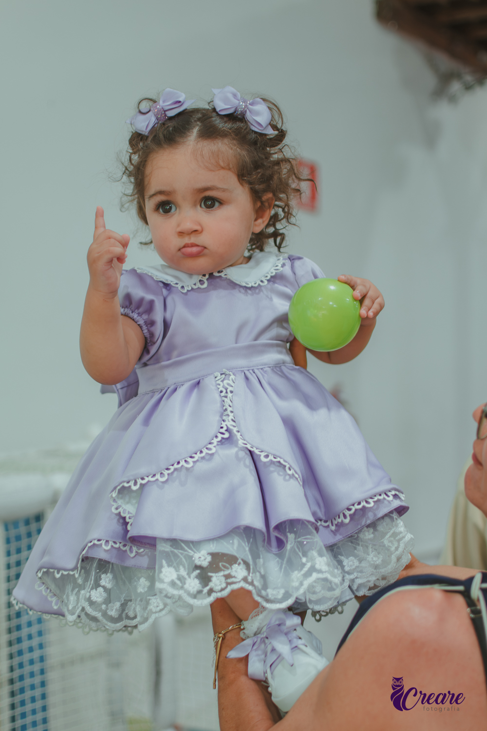 Uma bebê usando vestido roxo e segurando uma bolinha verde, durante seu aniversário de 1 ano.