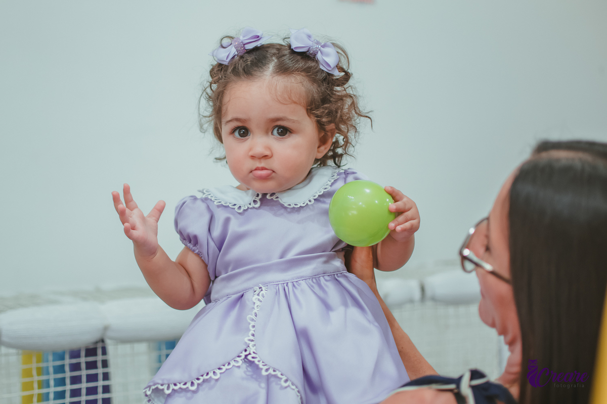 Uma bebê usando vestido roxo e segurando uma bolinha verde, durante seu aniversário de 1 ano.